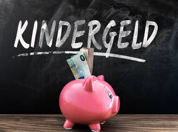 Слово KINDERGELD, немецкий для пособия на ребенка, на доске с поросячьим банком и наличными перед на деревянном столе — стоковое фото
