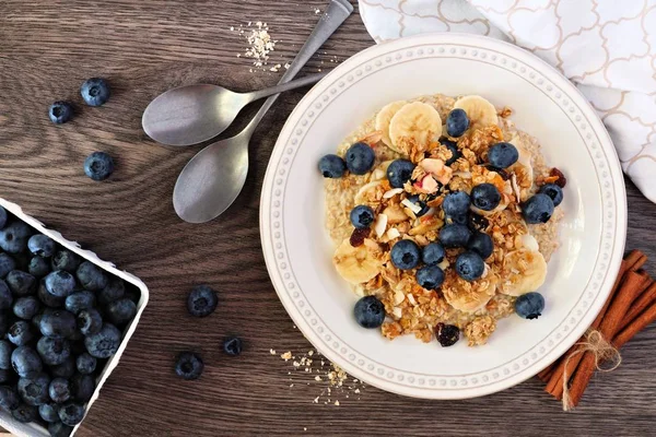 Healthy breakfast oatmeal topped with blueberries, bananas and granola. Table scene with a rustic wood background.