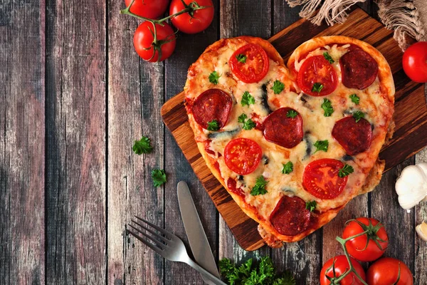 Heart shaped pizza for Valentines Day over a dark wood background. Top view, side border. Table scene with ingredients.