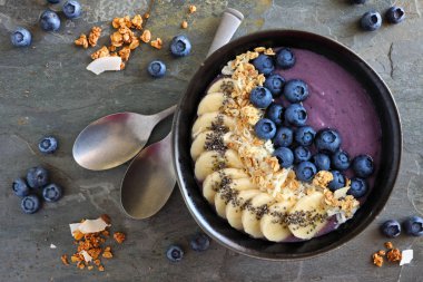 Hindistan cevizi, muz, chia tohum ve granola ile sağlıklı blueberry smoothie kase. Üstten görünüm tablo sahne karanlık bir taş zemin üzerine.