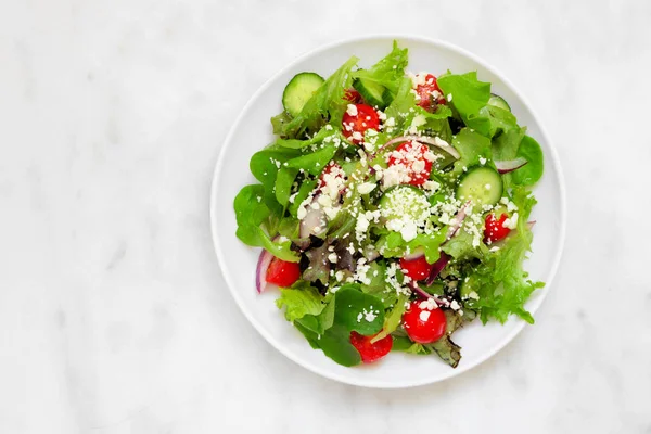 Healthy mixed salad with mixed greens, cucumber, onion, tomatoes and feta cheese. Top view against a white marble background.