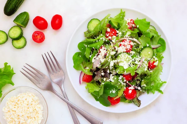 Healthy mixed salad with mixed greens, cucumber, onion, tomatoes and feta cheese. Top view table scene against a white marble background.