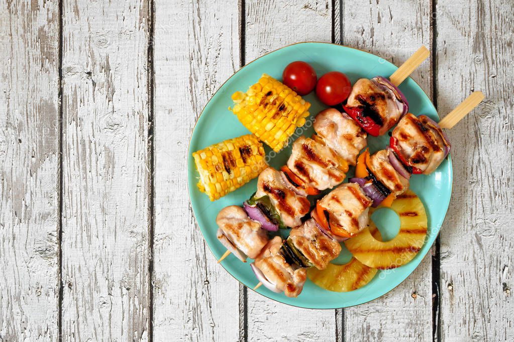 Plate of grilled chicken kabobs, corn, and pineapple. Top view on a white wood background. Summer BBQ or picnic food concept.