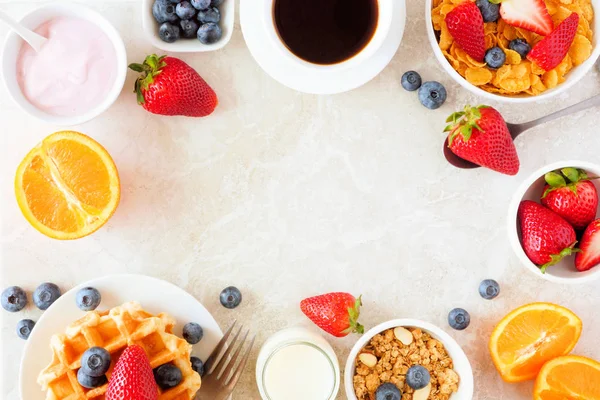 Breakfast food frame. Fruits, cereal, waffles, yogurt, coffee and milk. Top view over a bright stone background with copy space.