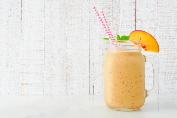 Peach smoothie in a mason jar glass with paper straws.  Side view with a white wood background.
