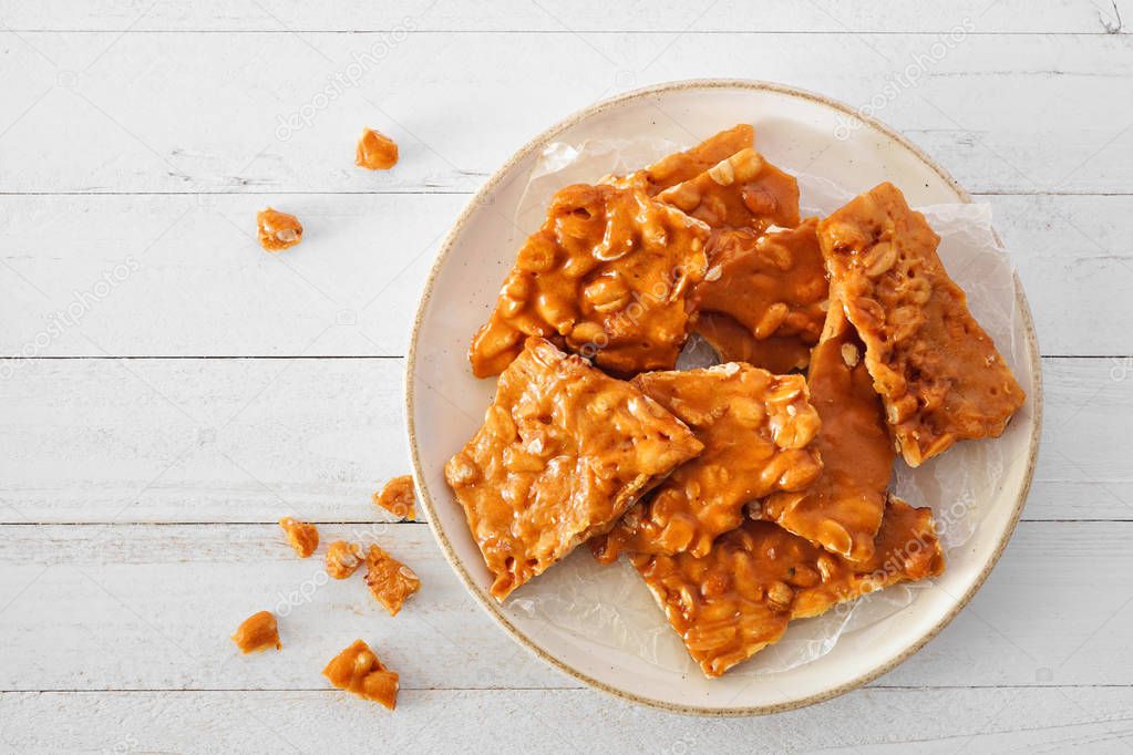 Plate of traditional peanut brittle candy pieces. Top view on a white wood background.