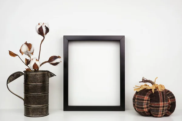 Mock up black frame with cotton branch and plaid pumpkin decor on a shelf or desk. Autumn concept. Portrait frame against a white wall.