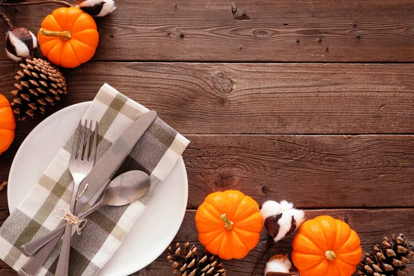 Fall corner border with dinner plate, silverware, check print napkin, pumpkins and pine cones. Autumn harvest or thanksgiving concept. Top view against a rustic wood background with copy space.