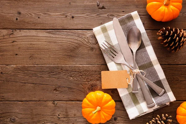 Blank gift tag with silverware, check print napkin, pumpkins and pine cones. Autumn harvest or thanksgiving concept. Top view side border against a rustic wood background with copy space.