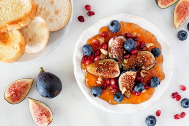 Baked brie appetizer garnished with figs and jam, blueberries, pomegranates and nuts. Top view scene on a white marble background.