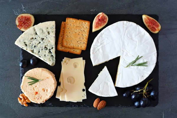 Cheese board with a selection of cheeses, crackers, figs and nuts on slate serving board. Above view on a dark background.