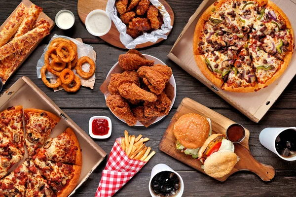 Büfe masası yemek servisi ya da teslimat sahnesi. Pizza, hamburger, kızarmış tavuk ve mezeler. Koyu bir ahşap arka planda manzaranın üstünde.