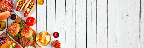 Summer BBQ food table scene with hot dog and hamburger buffet. Above view corner border over a white wood banner background. Copy space.