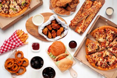 Büfe masası yemek servisi ya da teslimat sahnesi. Pizza, hamburger, kızarmış tavuk ve mezeler. Beyaz ahşap arka planda üst görünüm.