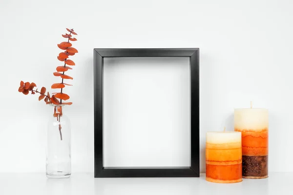 Mock up black frame with fall branches and candle decor on a white shelf. Autumn concept. Portrait frame against a white wall.