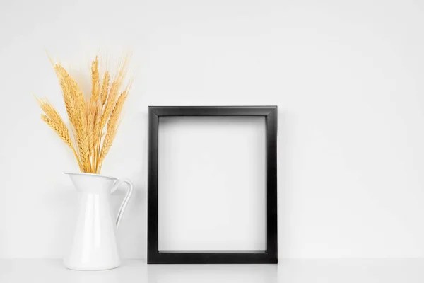 Mock up black frame with fall wheat in jug on a white shelf. Autumn concept. Portrait frame against a white wall.