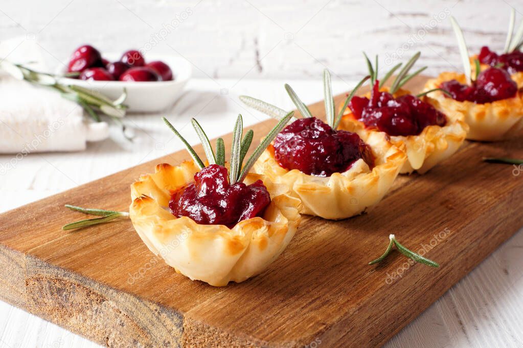 Holiday phyllo pastry appetizers with cranberries and baked brie. Serving board closeup against a white wood background.