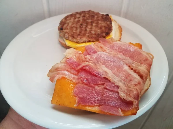 Tangan memegang sosis, telur, dan bacon dan keju sandwich di piring putih — Stok Foto