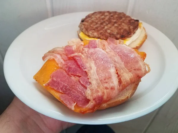 Mão segurando salsicha, ovo, bacon e queijo sanduíche no prato branco — Fotografia de Stock