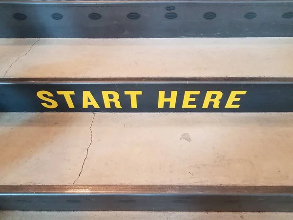 yellow start here sign on black and grey cement stairs or steps