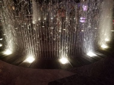 İnsanlar geceleri dışarıdaki ışıklarla çeşme jetlerinde oynuyorlar.