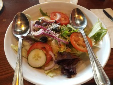 Salatalı, salatalı, domatesli ve kaşıklı beyaz bir kase.