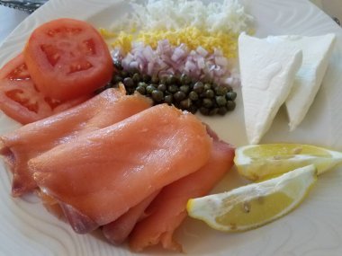 Tütsülenmiş somon, limon, domates ve kapari dolu beyaz bir tabak.