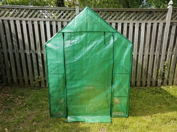 green greenhouse with door on grass with wood fence