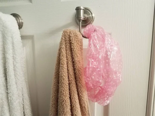白色和棕色的毛巾挂在浴室门上的钩子上 上面有粉色的浴帽 — 图库照片