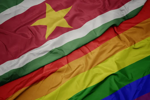 Waving colorful gay rainbow flag and national flag of suriname. — Stock Photo, Image