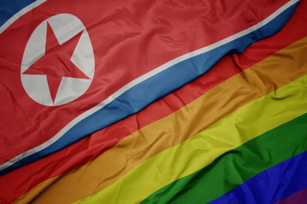 Hullámzó színes meleg szivárványos zászló és a nemzeti zászló Észak-Korea. — Stock Fotó