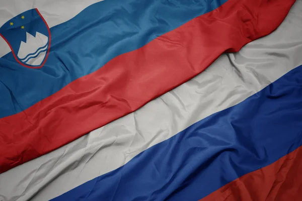 Mávající barvitá vlajka Ruska a národní vlajka Slovinska. — Stock fotografie