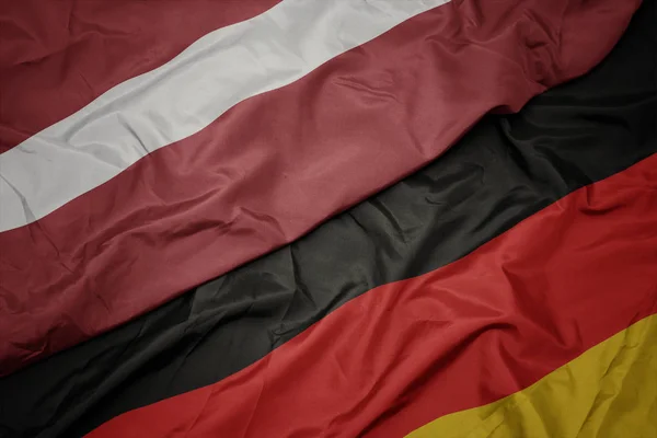 Mávající barvitá vlajka Německa a národní vlajka Lotyšska. — Stock fotografie