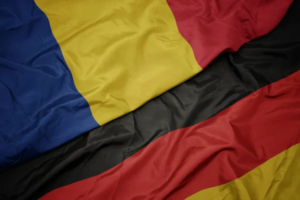 Zwaaiende kleurrijke vlag van Duitsland en de nationale vlag van Roemenië. — Stockfoto