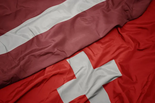 Falujące kolorowe flagi Szwajcarii i flagi narodowej Łotwy. — Zdjęcie stockowe