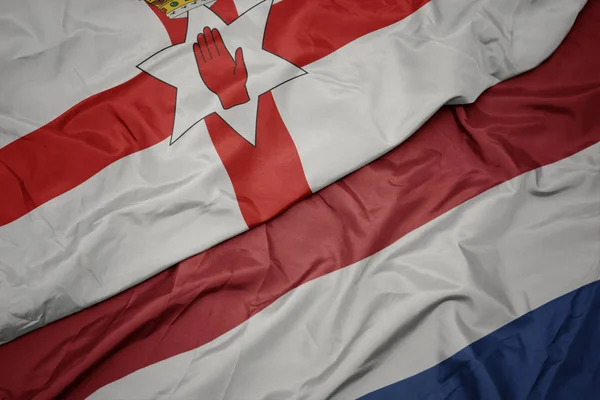 Ondeando colorida bandera de los Países Bajos y bandera nacional de Irlanda del Norte . — Foto de Stock