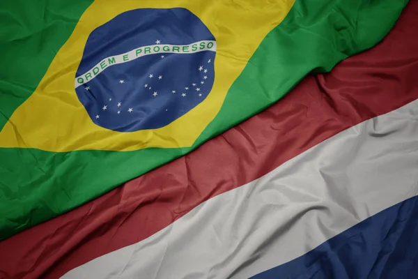 Ondeando colorida bandera de los Países Bajos y bandera nacional de Brasil . — Foto de Stock