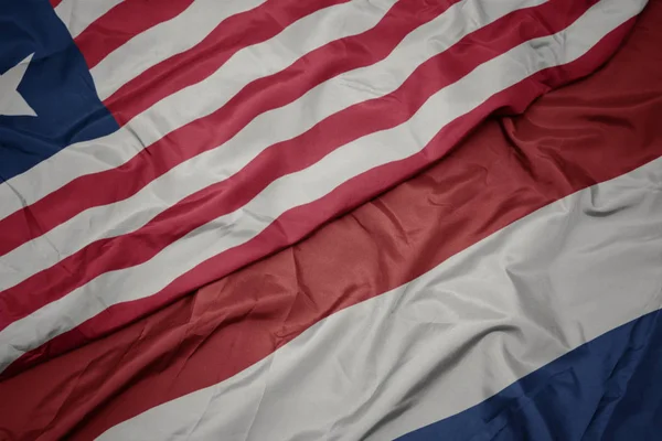 Ondeando colorida bandera de los Países Bajos y bandera nacional de Liberia . — Foto de Stock