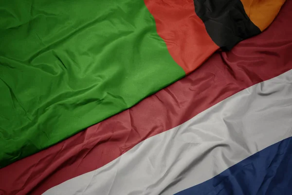 Mávající barevná vlajka Nizozemska a národní vlajka Zambie. — Stock fotografie