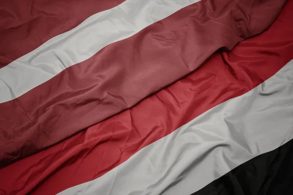 Zwaaiende kleurrijke vlag van Jemen en de nationale vlag van Letland. — Stockfoto