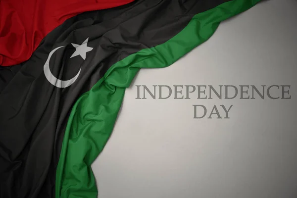 Machając kolorowe flagi narodowej Libii na szarym tle z dniem niepodległości tekstu. — Zdjęcie stockowe