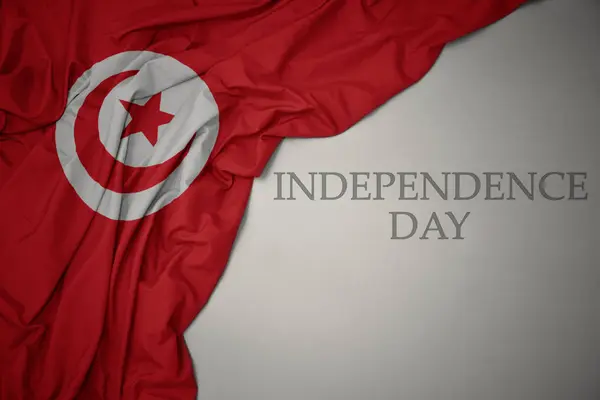 Machając kolorowe flagi narodowej Tunezji na szarym tle z dniem niepodległości tekstu. — Zdjęcie stockowe
