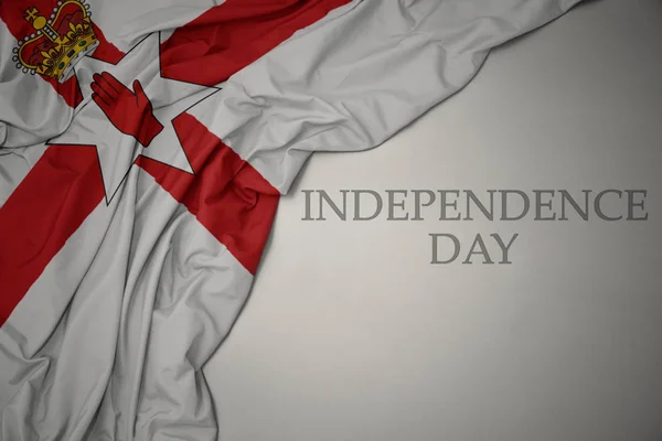 Zwaaiende kleurrijke nationale vlag van Noord-Ierland op een grijze achtergrond met tekst Onafhankelijkheidsdag. — Stockfoto
