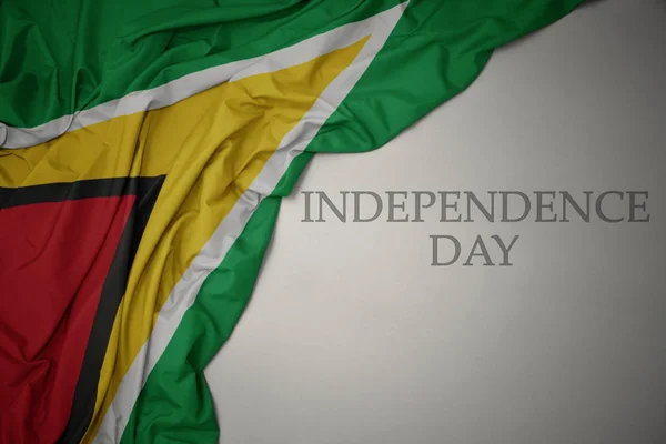 Zwaaiende kleurrijke nationale vlag van Guyana op een grijze achtergrond met tekst Independence Day. — Stockfoto