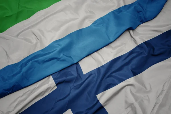 Ondeando colorida bandera de Finlandia y bandera nacional de Sierra Leona . — Foto de Stock