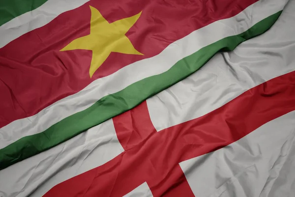 Ondeando colorida bandera de Inglaterra y bandera nacional de Surinam . — Foto de Stock