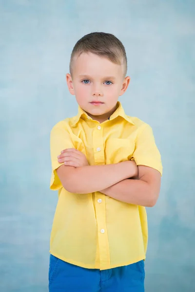 Cute confident serious caucasian little boy in a yellow shirt with arms crossed on a blue background — Stock Photo, Image