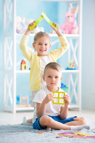 A little girl with a boy is sitting on the floor and holding a house of colored paper. — Stock Photo, Image