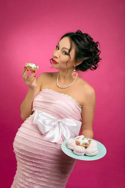 pregnant girl in pin-up style with a cake in a pink dress on a pink background.