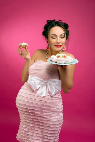 pregnant girl in pin-up style with a cake in a pink dress on a pink background.